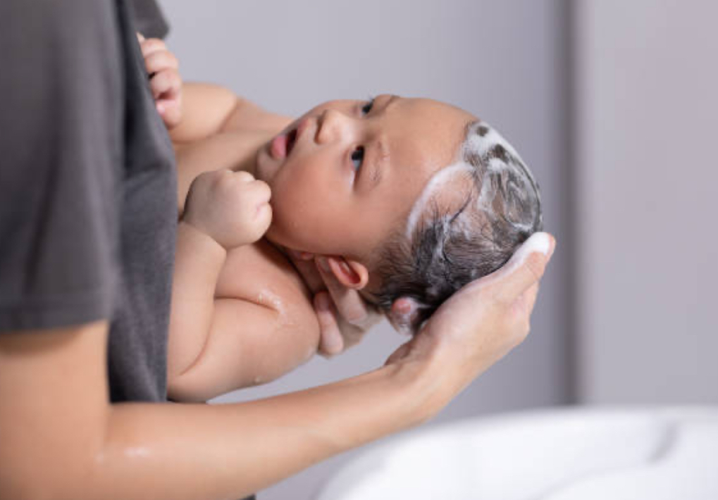 A parent's hands gently washing a baby's hair with foam, focusing on the tender, careful touch